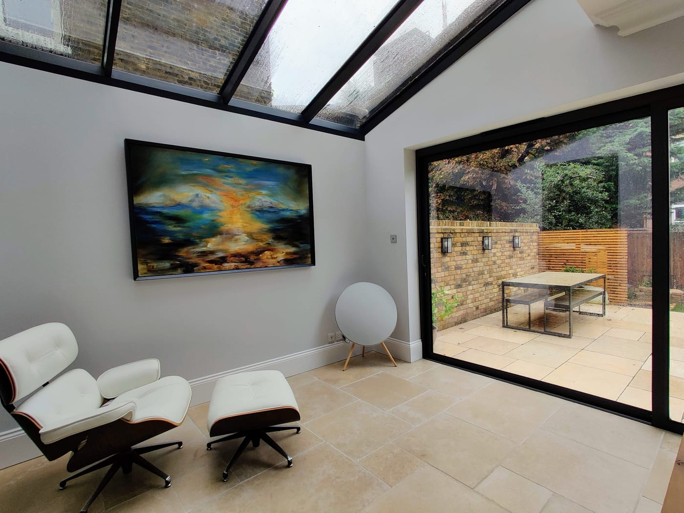 A wide abstract landscape in blues and gold hung in a client's glass-roofed kitchen extension, a white lounge chair in front and the garden beyond