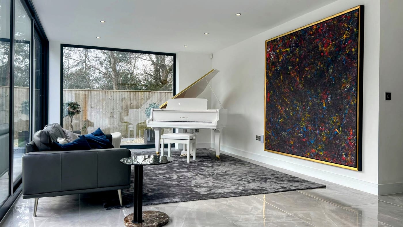 A very large gold-framed abstract, flecks of red, yellow and blue on black, on the white wall of a client's garden room beside a white grand piano