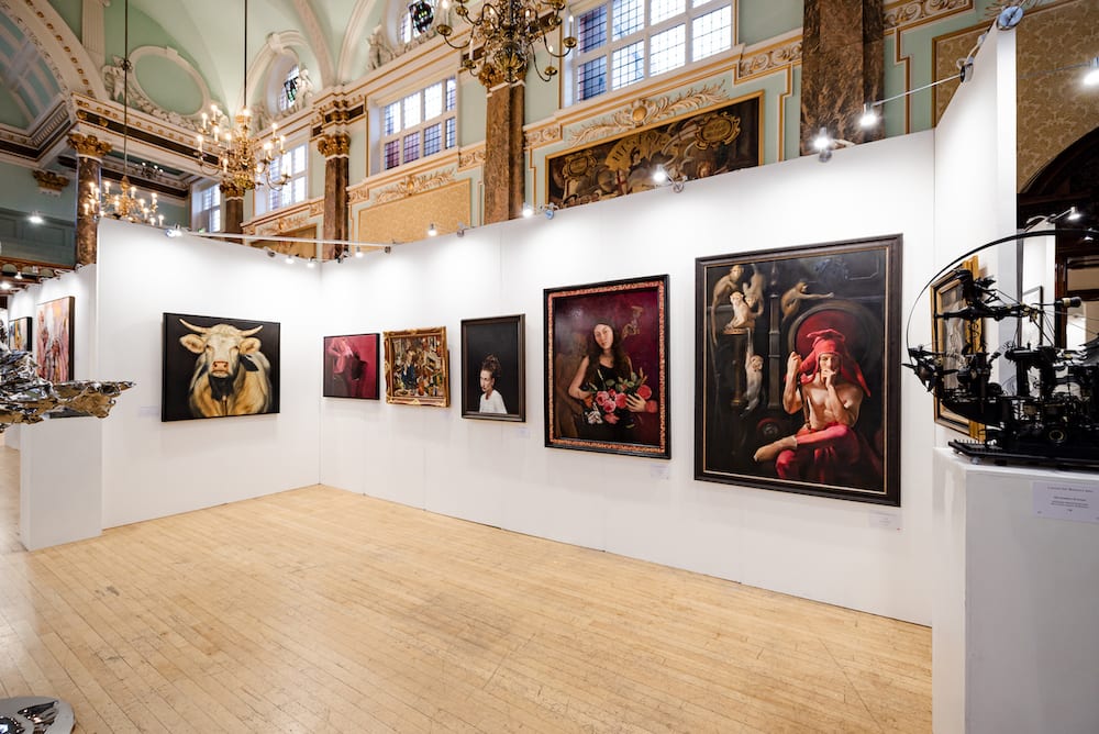 The London Art Biennale hang at Chelsea Old Town Hall — portraits and paintings on white walls beneath the hall's painted ceiling